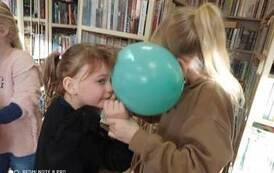 Para dziewczynek trzymających się rękami twarzą do siebie i głowami trzymają wsp&oacute;lnie zielony balon 