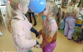 Dwie dziewczynki w parze trzymają się za ręce i z twarzą do siebie trzymają głowami balon.