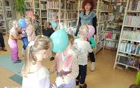 Dzieci tanczą w parach trzymając się za ręce i głowami trzymając wsp&oacute;lnie balonik. Widać Panią Nauczycielkę., kt&oacute;ra stoi i patrzy.