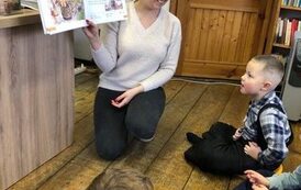 Dzieci siedzą na poduszkach na podłodze i patrzą na bibliotekarkę, kt&oacute;ra czyta i pokazuje im  książki o tematyce kociewskiej.