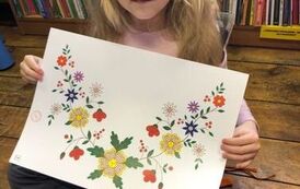 Dziewczynka o blond włosach siedzi na podłodze, w dłoniach trzyma dużą kartkę z wydrokowanym haftem kociewskim.