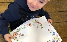 Chłopiec siedzi na podłodze, trzyma w dłoniach dużą kartke na kt&oacute;rej jest narysowany haft kociewski.