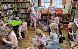 Dzieci siedzą na podłodze i krzesełkach patrząc na koleżankę,kt&oacute;ra stoi przy p&oacute;łce z książeczkami na kt&oacute;rych są obrazki Czerwonego Kapturka.