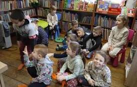 Dzieci siedzą w rozsypce na poduszkach i krzesełkach słuchając czytanej bajki.