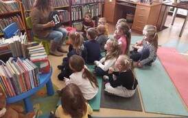Dzieci siedzą na karimatach i patrzą na bibliotekarkę, kt&oacute;ra im pokazuje czarne, okrągłe siedzisko z numerem 10.