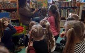Dzieci siedzą na karimatach,a bibliotekarka czyta im bajkę w formacie 3D o dinozaurach pokazując przy tym ilustracje.
