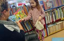 Bibliotekarka pokazuje książeczkę pluszową, a przy Niej stoi dziewczynka z siateczką aby zabawić się w robienie zakup&oacute;w z książeczki. Rączką odczepia z książeczki jakiś przedmiot.