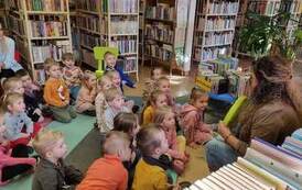 Dzieci siedzą na karimatach,a bibliotekarka pokazuje im zielone siedzisko. Z tyłu widać na krzesełku opiekunkę.