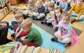 Dzieci siedzą na karimatach słuchając czytanej bajki przez bibliotekarkę.