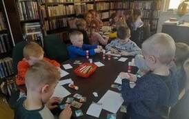 Przy dw&oacute;ch dużych stołach siedzą dzieci i naklejają na białą kartkę puzzle z bajki "Trzy małe świnki".