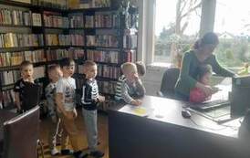 Dzieci stoją w kolejce przy biurku aby skorzystać z czytnika,a bibliotekarka pomaga dziecku przyłożyć książkę do czytanika.