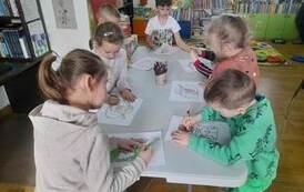 Dzieci stoją przy białym stole i  kolorują postaci z baśni Andersena.