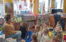 Dzieci siedzą na dywanie i karimatach skierowane do bibliotekarki czytającej im bajkę.