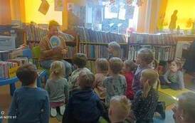 Dzieci siedzą na dywanie i karimatach skierowane do bibliotekarki, kt&oacute;ra z uśmiechem pokazuje im okładkę tytułową wielkanocnej książeczki w kształcie jajka.