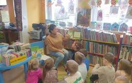 Na dywanie siedzą dzieci i słuchają bajki czytanej przez bibliotekarkę. 