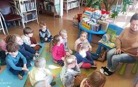 Dzieci siedzą na dywanie i karimatach skierowane do bibliotekarki czytającej im bajkę.