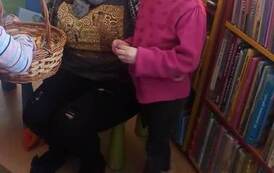 Mała dziewczynka o blond włosach stoi przy bibliotekarce, kt&oacute;ra trzyma koszyk z cukierkami. W koszyku widać czyjąś rączkę.