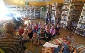 Dzieci siedzą na dywanie, a bibliotekarka czyta im bajkę.Z tyłu siedzi na krześle opiekenka dzieci.