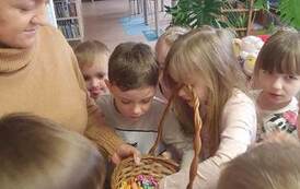 Bibliotekarka stoi z koszykiem cukierk&oacute;w otoczona częstującymi się dziećmi.