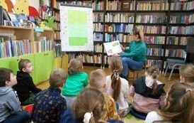 Dzieci siedzą na dywanie i słuchają bajki czytanej przez bibliotekarkę, po lewej stronie widać metalową tablicę z przyczepioną do niej krzyż&oacute;wką do rozwiązania.