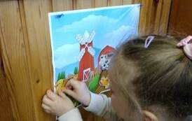 Na drzwiach jest przyczepiony plakat na kt&oacute;rym jest farma i dziewczynka przykleja na nim zwierzątko.