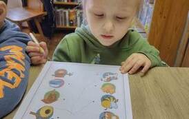 Dziewczynka stoi przy stoliku i na kartce rysując łączy zwierzątka z ich domkami.