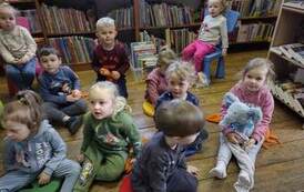 Dzieci siedzą na poduszkach i słuchają bajki czytanej przez bibliotekarkę.