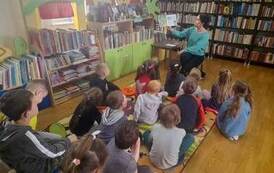 Dzieci siedzą na dywanie, a bibliotekarka siedzi przed nimi i czyta im bajkę.