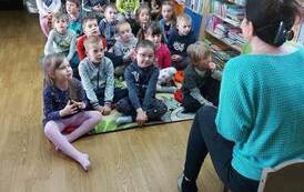 Dzieci siedzą na dywanie i patrzą na bibliotekarkę, kt&oacute;ra siedzi z przodu i coś do nich m&oacute;wi.