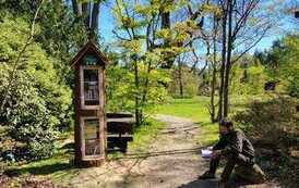 Mężczyzna siedzi na kamieniu przy biblioteczce w Wirtach czytając książkę.