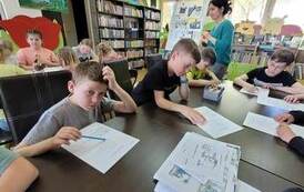 Dzieci siedzą przy stołach i rozwiązują krzyż&oacute;wkę, na stole są poprzednie prace zrobione przez dzieci. w tle widac bibliotekarkę.