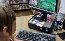 Dziewczynka stoi przy biurku przy komputerze rozwiązując zagadki, kt&oacute;re pojawiają sie na monitorze.