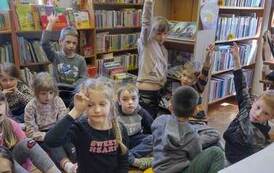 Dzieci siedzą na siedziskach i słuchają bibliotekarza. Niekt&oacute;re z dzieci podnoszą rękę zgłaszając się.