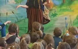 Na leśnym tle stoi aktorka przebrana za dziewczynkę, z tyłu ma plecak i m&oacute;wi do małych widz&oacute;w.