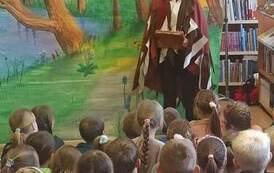 Z przodu stoi przebrany aktor i trzyma książkę,przed Nim siedzą dzieci i patrzą. W tle widać las. 