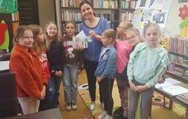 Bibliotekarka stoi z paroma dziewczynkami trzymając od nich laurki i świeczkę. Bardzo się uśmiecha.