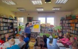 Dzieci siedzą przy stołach i patrzą do przodu na bibliotekarkę, kt&oacute;ra przyczepia do metalowej tablicy ilustrację dotyczącą powstawania książki.