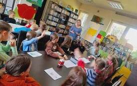 Dzieci siedzą przy stołach i patrzą w stronę bibliotekarki, kt&oacute;ra m&oacute;wi im o powstawaniu książki.