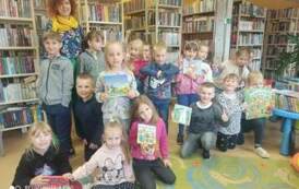Pani wychowawczyni stoi ze swoimi dziećmi w grupie w bibliotece. Niekt&oacute;re z dzieci trzymają książeczki bajeczki.
