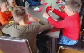 Dzieci siedzą przy stołach i wycinają z czerwonej bibuły płatki do kwiata.