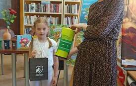 Pani Gosia stoi i trzyma zgniatarkę do butelek, a z lewej strony stoi dziewczynka z czarną torebką, w kt&oacute;rej jest upominek dla Pani Gosi za czytanie.