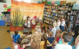 Z przodu siedzi indianka z walizką i trzyma w ręku misia z r&oacute;żowym kapeluszem.W tle widać makietę przedstawiająca prerię. Widownia patrzy z zainteresowaniem, jeden z chłopc&oacute;w stoi.