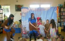 Kilkoro dzieci tańczy z przodu w k&oacute;łeczku z Indianką. Obok dzieci siedzących tańczy w długich czarnych włosach Induanin z niebieskimi frędzlami.