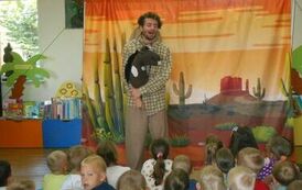 Aktor w koszuli w kratę i beżowych spodniach trzyma w ręku konika na kiju. Widać publiczność dziecięcą, a w tle makieta przedstawiająca prerię.