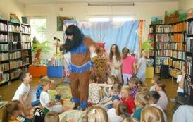 Aktor w czarnych długich włosach przebrany za indianina jest wśr&oacute;d dzieci i tańczy.