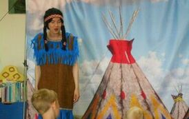 Aktorka przebrana za indiankę w warkoczykach stoi i m&oacute;wi coś do dzieci. W tle widać makietę z namiotem i dziecko z widowni.