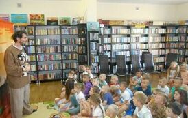 Duża grupa dzieci siedzi na dywanie,a z przodu stoi aktor teatrzyku i coś do nich m&oacute;wi. 