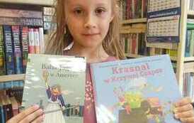 Dziewczynka o blond włosach trzyma wypożyczoną książkę i książkę z wyprawki czytelniczej.