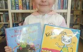 Dziewczynka o blond włosach trzyma wypożyczoną książkę i książkę z wyprawki czytelniczej.
