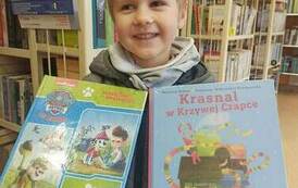 Chłopiec o bujnych blond wlosach stoi uśmiechnięty i trzyma dwie książki : wypożyczoną i i z wyprawki.